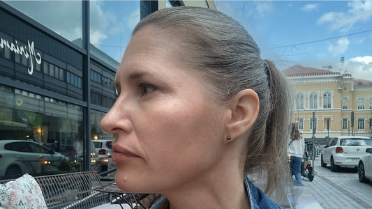 Sivuprofiilikuva Katjasta - naisesta, joka istuu ulkona kahvilan terassilla kaupunkimaisemassa. Taustalla heijastuu rakennuksia ja ohikulkevia ihmisiä. Katja miettii, mikseivät apuvälineet voisi olla kateuden kohteita.