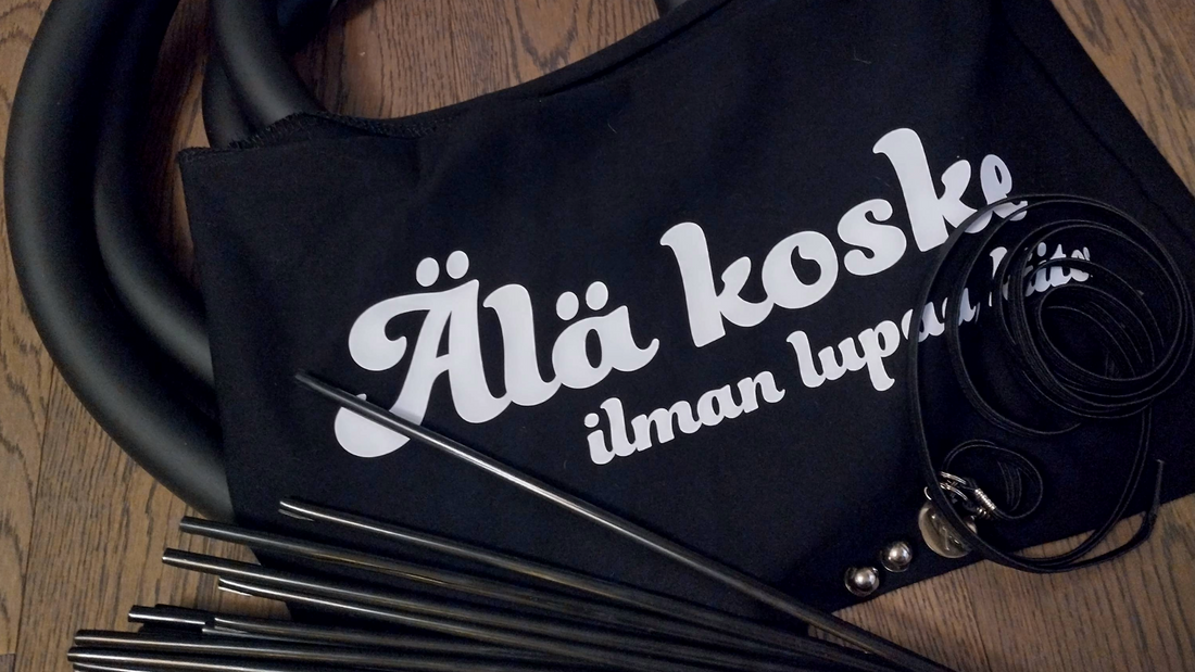 Tummalla puualustalla mustat kelausvanteen suojat, mustat pinnakuoret ja mustat kengännauhat, sekä musta pyörätuoli-bandeau, jossa valkoinen teksti: "älä koske ilman lupaa, kiitos."